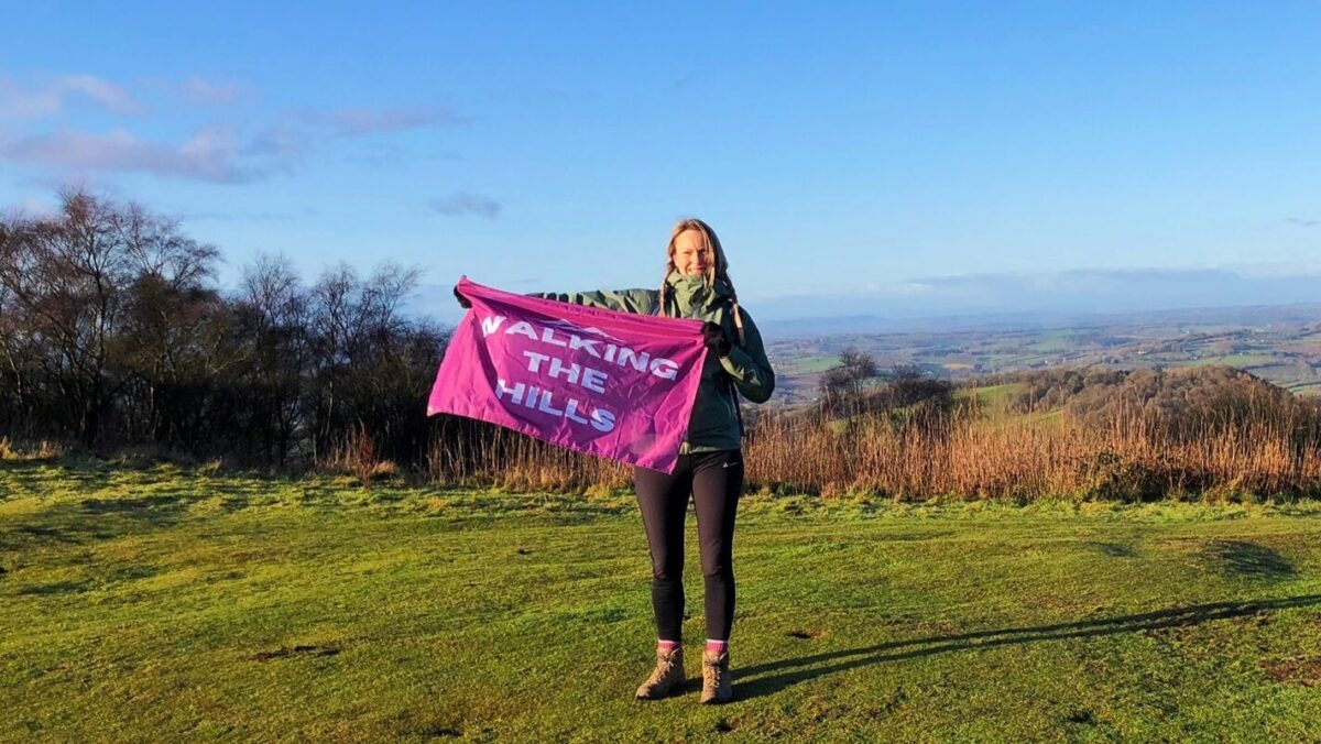 A woman standing on the hills holding a pink flag with the text 'Walking the Hills'