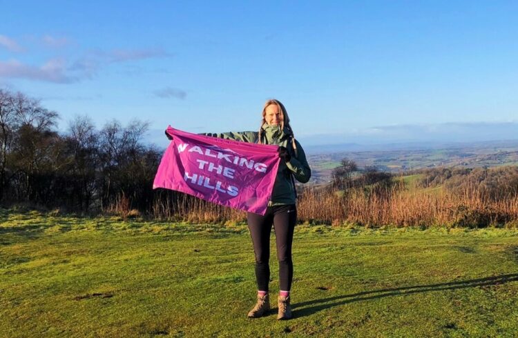 A woman standing on the hills holding a pink flag with the text 'Walking the Hills'